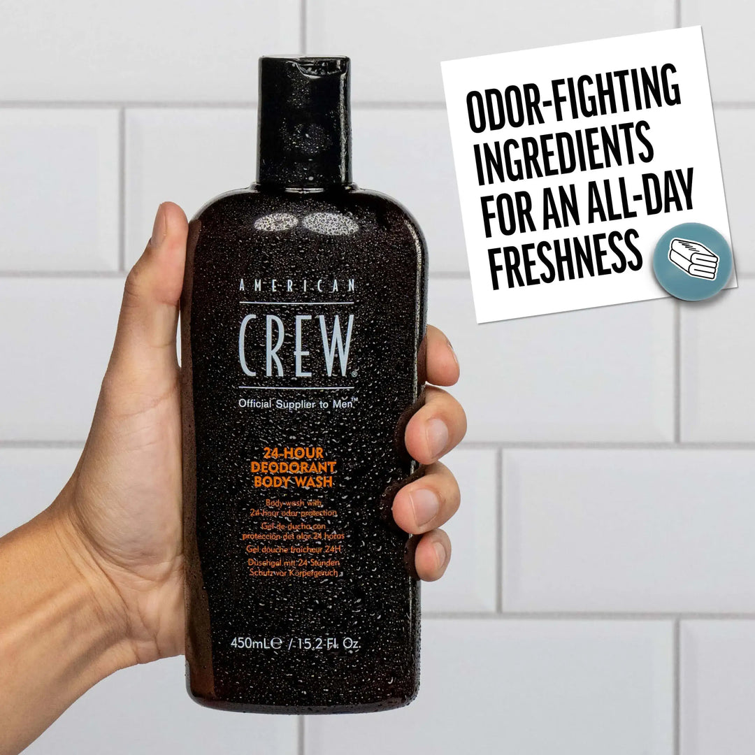 Hand holding a bottle of American Crew 24 Hour Deodorant Body Wash with a tiled bathroom background. Callout states "Odor-fighting ingredients for an all-day freshness". 