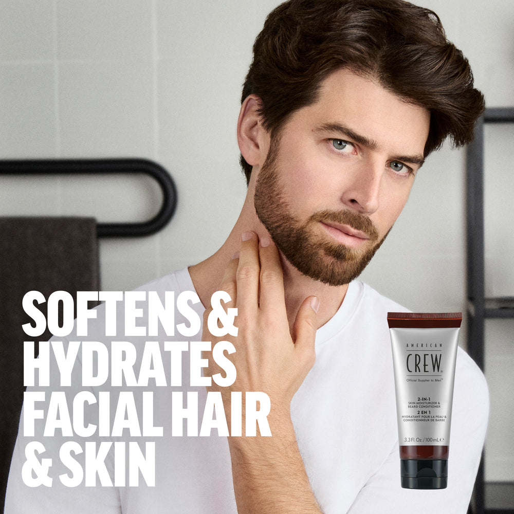 Model Ferrari Calderon is sporting a full beard and mustache holding his chin, while wearing a white shirt with an overlay of American Crew 2-IN-1 Skin Moisturizer and Beard Conditioner next to the text "Softens & hydrates facial hair & skin". 