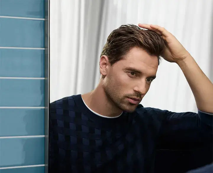 Ezrak, with styled brown hair looks over his shoulder while touching his hair, wearing a dark navy textured knit shirt with a white-collar trim. The background features a blue tiled wall on the left and white curtains behind him.