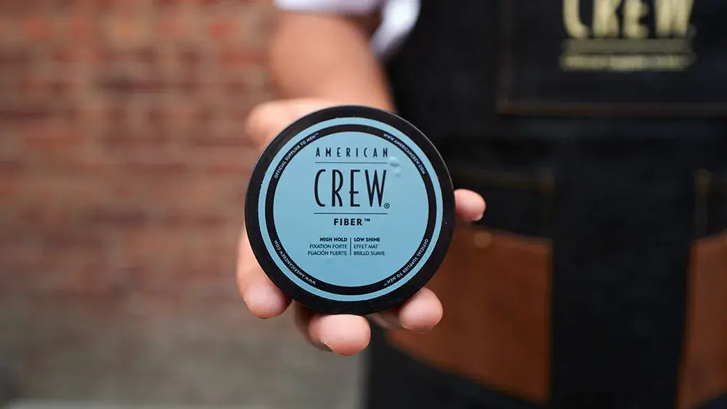 A hand holding a round black container of American Crew Fiber hair product against a blurred brick wall and dark apron background.