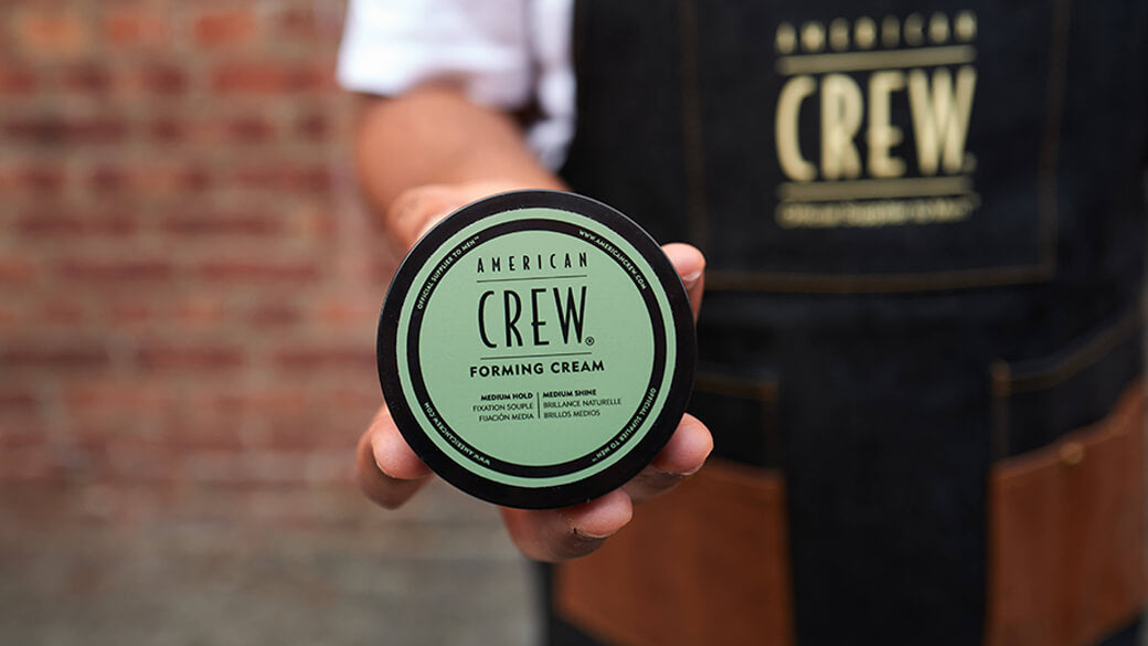A hand holding a round black container of American Crew Forming Cream, with a blurred brick wall and a person wearing a matching American Crew apron in the background.