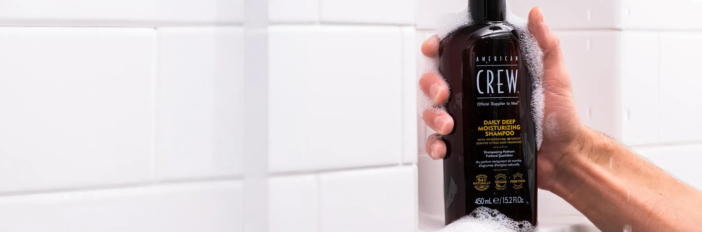 Man holding American Crew Daily Deep Moisturizing Shampoo in a shower cubby with soapy suds forming around the bottle. 