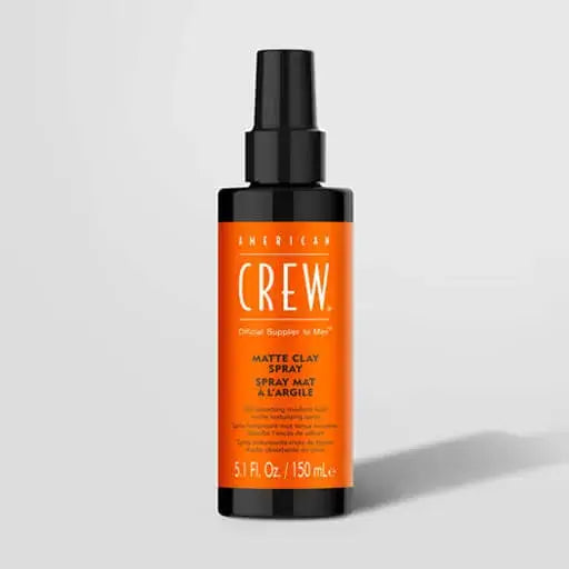 A bottle of American Crew Matte Clay Spray on a light gray background. The bottle is black with a prominent orange label and a black spray nozzle.