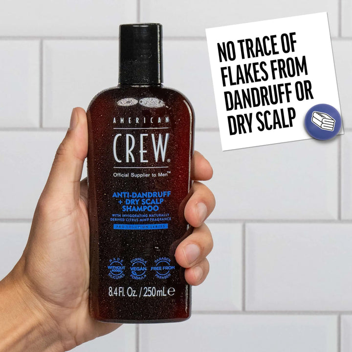 Hand holding a bottle of American Crew Anti-Dandruff + Dry Scalp Shampoo with a tiled bathroom background. Callout states "No Trace of Flakes from Dandruff or Dry Scalp". 