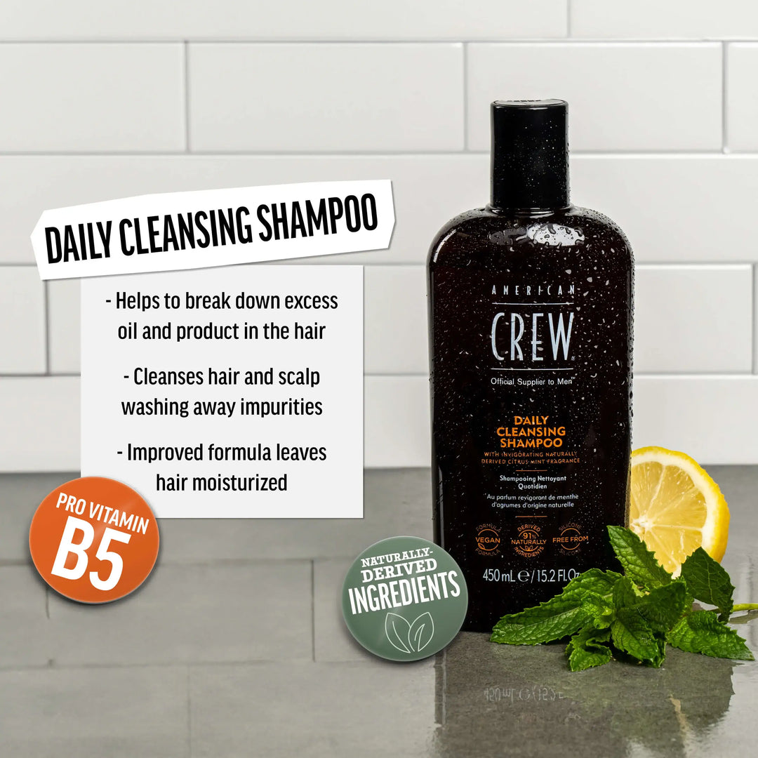 American Crew Daily Cleansing Shampoo bottle with lemon and mint leaves on a tiled background. Text callout says "Helps to break down excess oil and product in the hair. Cleanses hair and scalp washing away impurities. Improved formula leaves hair moisturized". 
