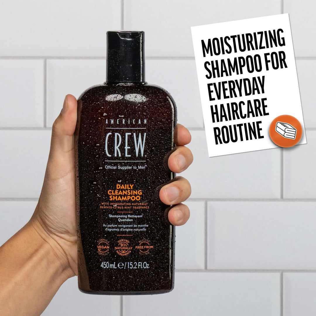 Hand holding a bottle of American Crew Daily Cleansing Shampoo with a tiled bathroom background. Callout states "Moisturizing Shampoo for Everyday Haircare Routine". 