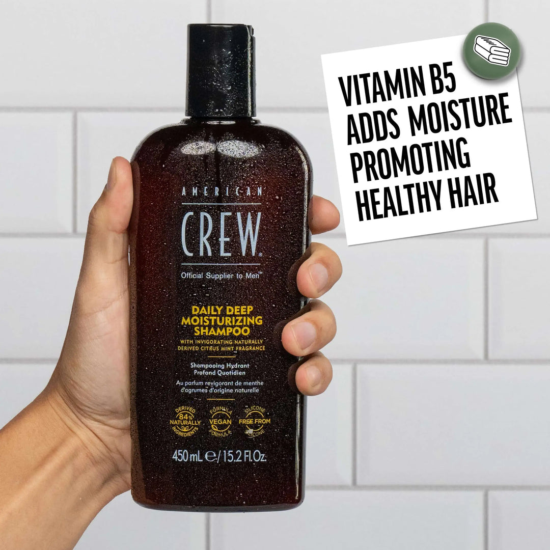 Hand holding a bottle of American Crew Daily Deep Moisturizing Shampoo with a tiled bathroom background. Callout states "Vitamin B5 Adds Moisture Promoting Healthy Hair". 