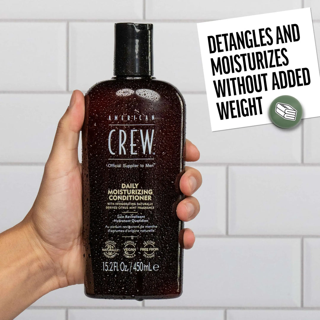 Hand holding a bottle of American Crew Daily Moisturizing Conditioner with a tiled bathroom background. Callout states "Detangles and Moisturizes without added weight". 