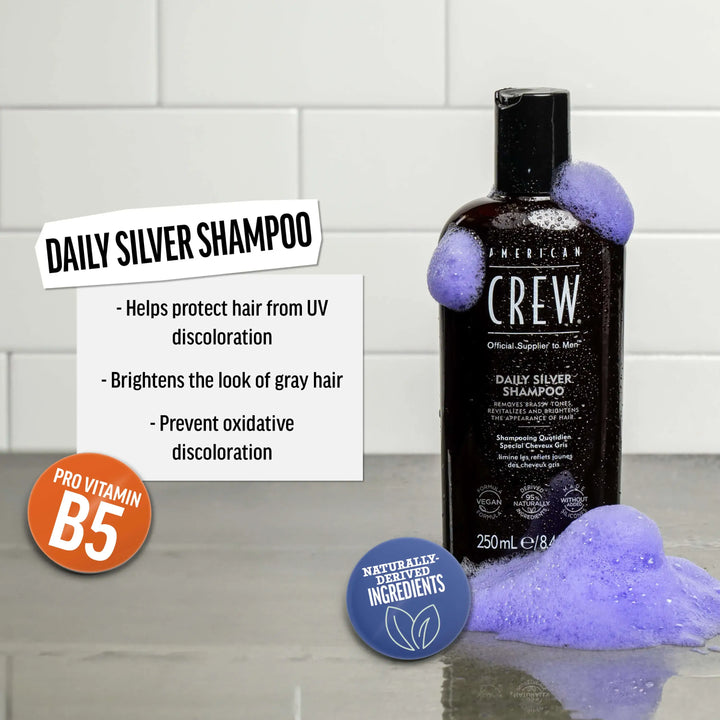 American Crew Daily Silver Shampoo on a bathroom shelf against a tiled background. Text callout says "Helps protect hair from UV discoloration. Brightens the look of gray hair. Prevents oxidative discoloration". 