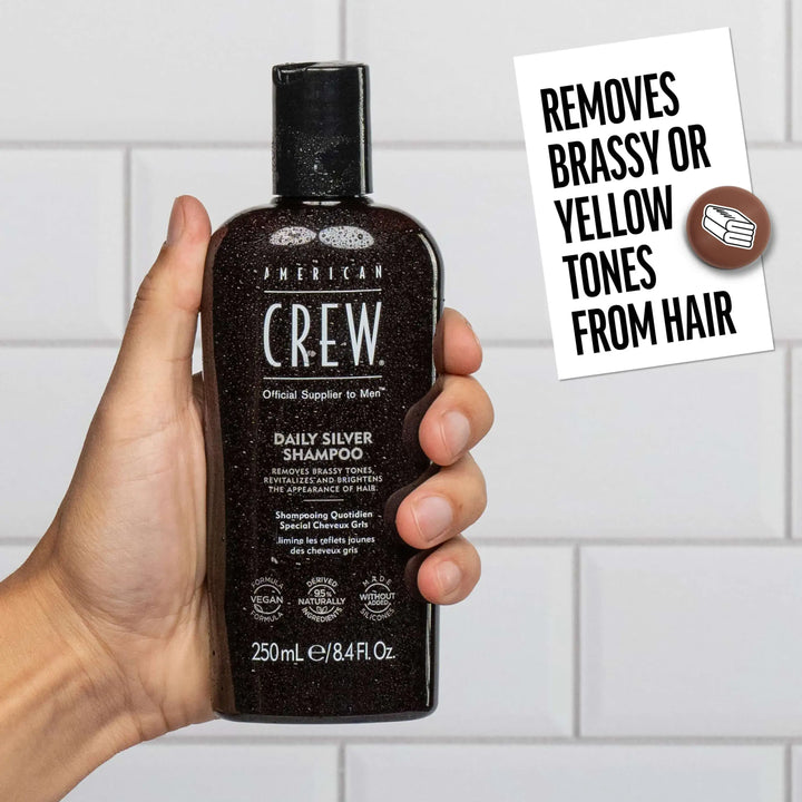 Hand holding a bottle of American Crew Silver Shampoo with a tiled bathroom background. Callout states "Removes brassy or yellow tones from hair". 