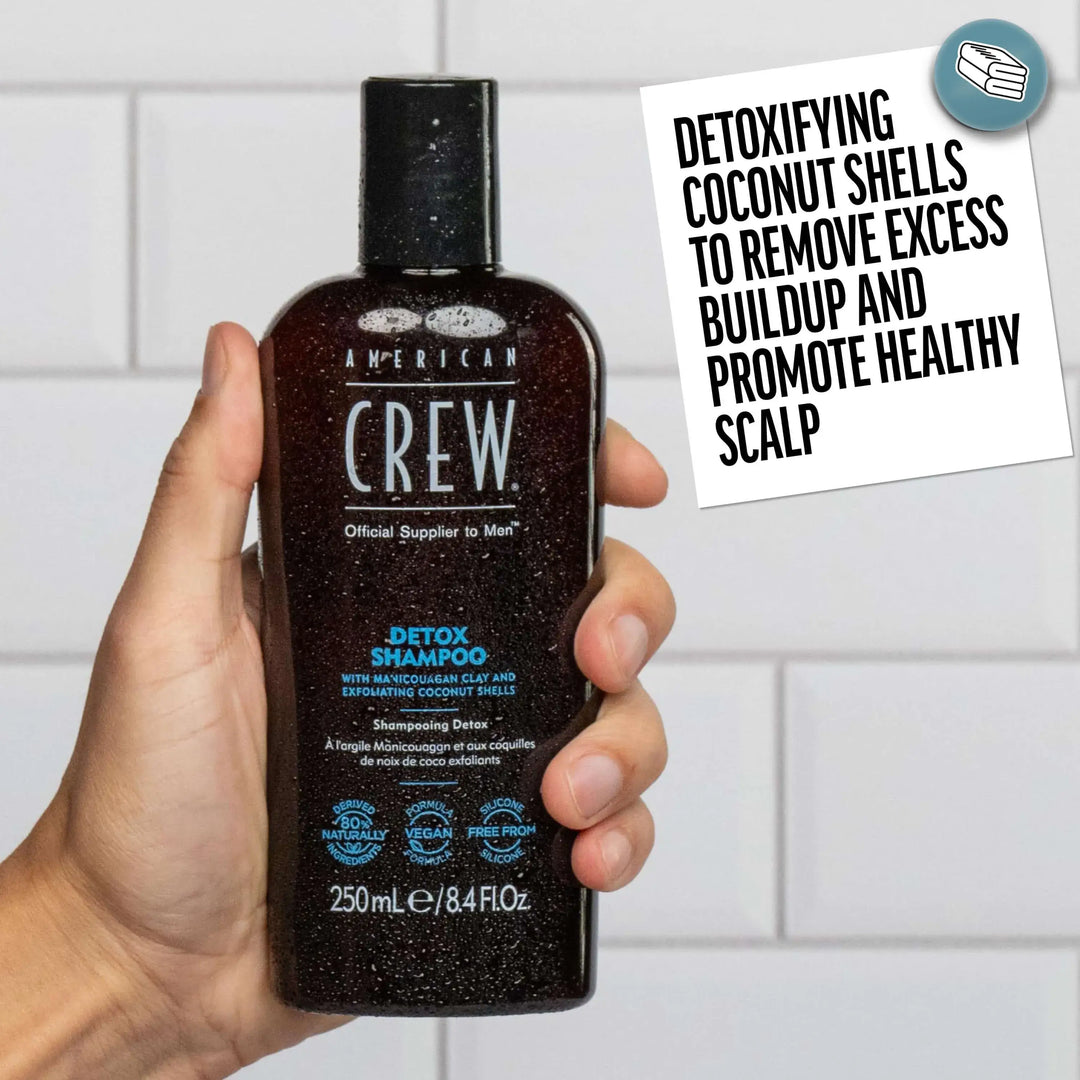 Hand holding a bottle of American Crew Daily Detox Shampoo with a tiled bathroom background. Callout states "Detoxifying Coconut Shells to remove excess bulid-up and promote healthy scalp". 